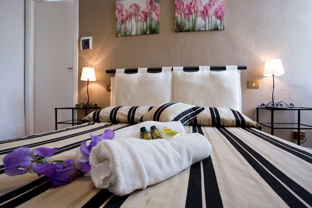 Quarto de hotel com uma cama, duas mesas de cabeceira, um abajur, uma toalha e um buquê de flores roxas.