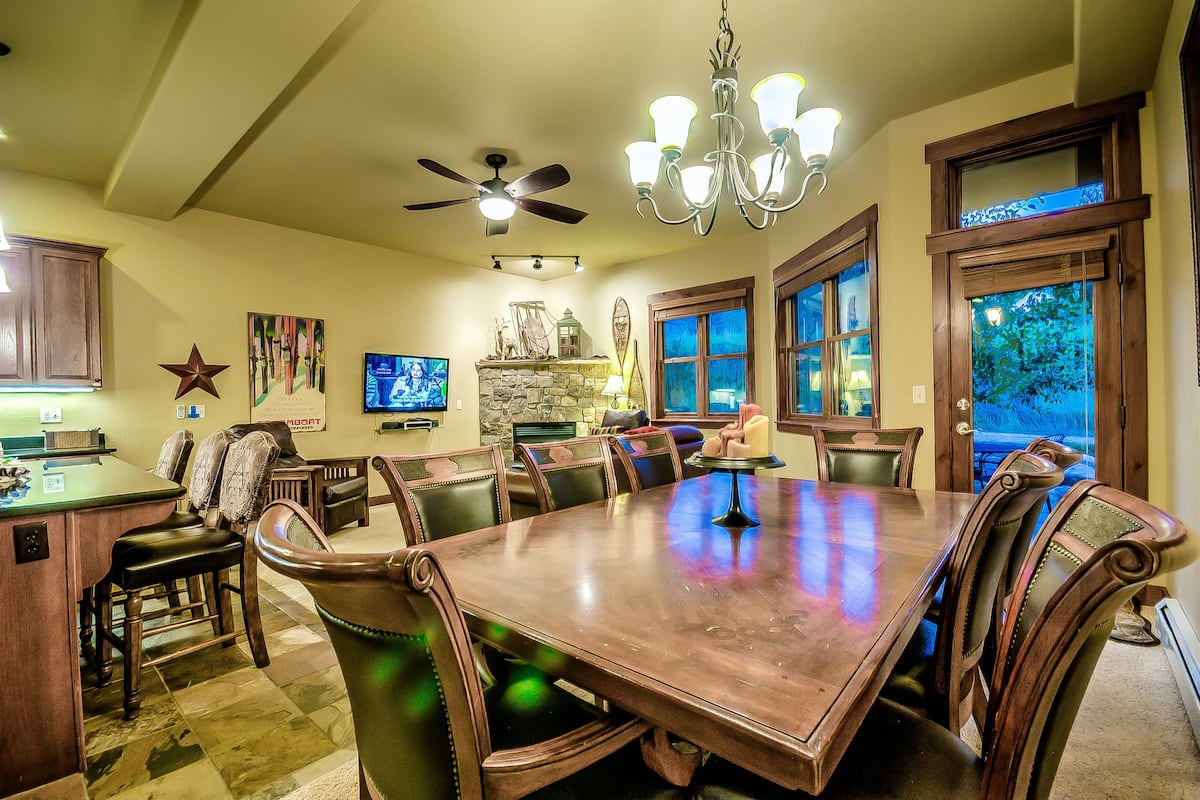 Une salle à manger avec une grande table en bois, plusieurs chaises, un lustre, un ventilateur de plafond et une cheminée en pierre.