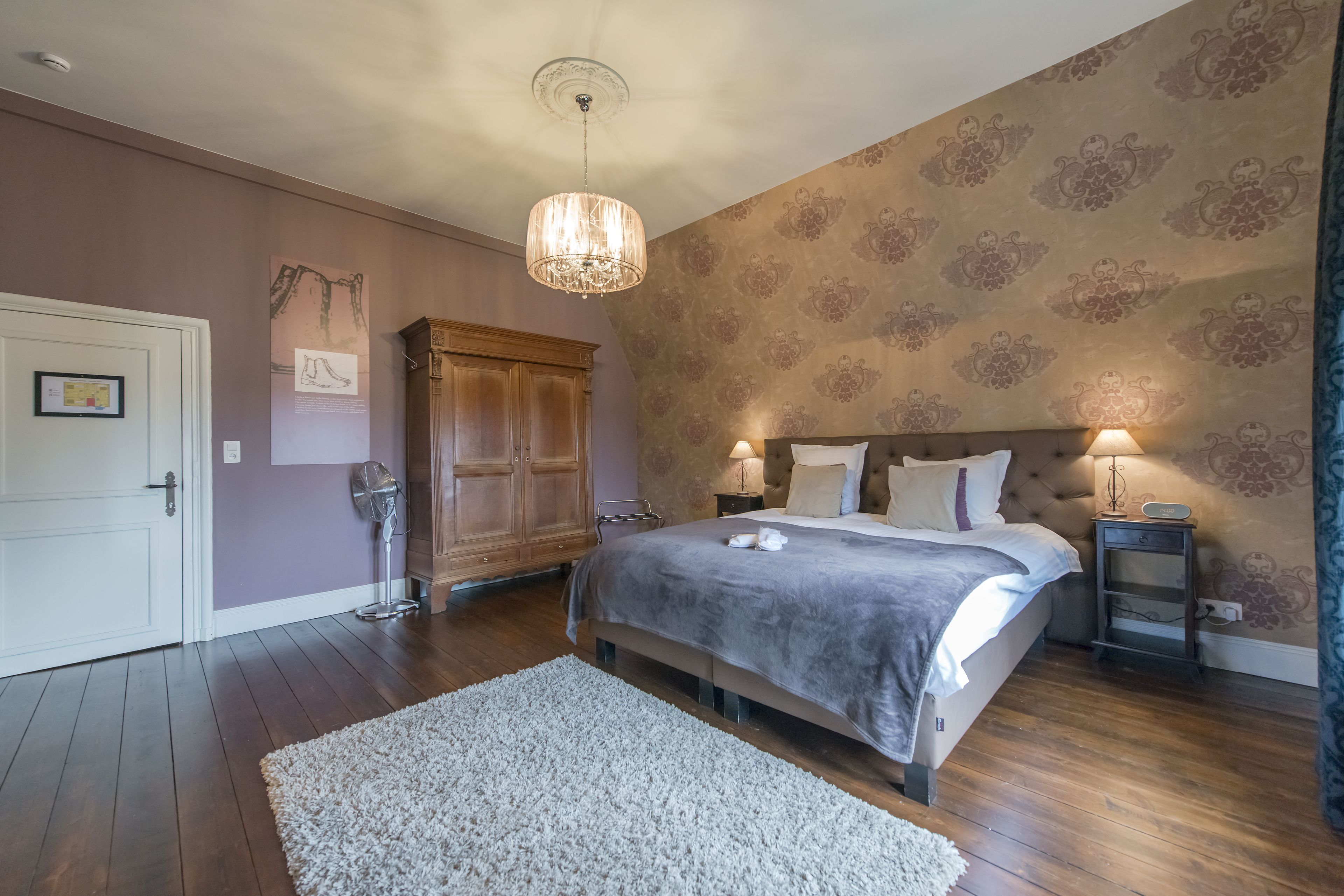 A bedroom with a large bed, a chandelier, a wooden wardrobe, and a patterned wallpaper.