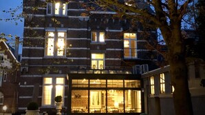 A traditional brick building with multiple windows and a large glass entrance, surrounded by trees and a well-maintained lawn. - 't Huys van Steyns (Tongeren)