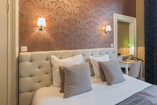 A hotel room with a tufted headboard, two bedside tables, a desk with a lamp, and a mirror.