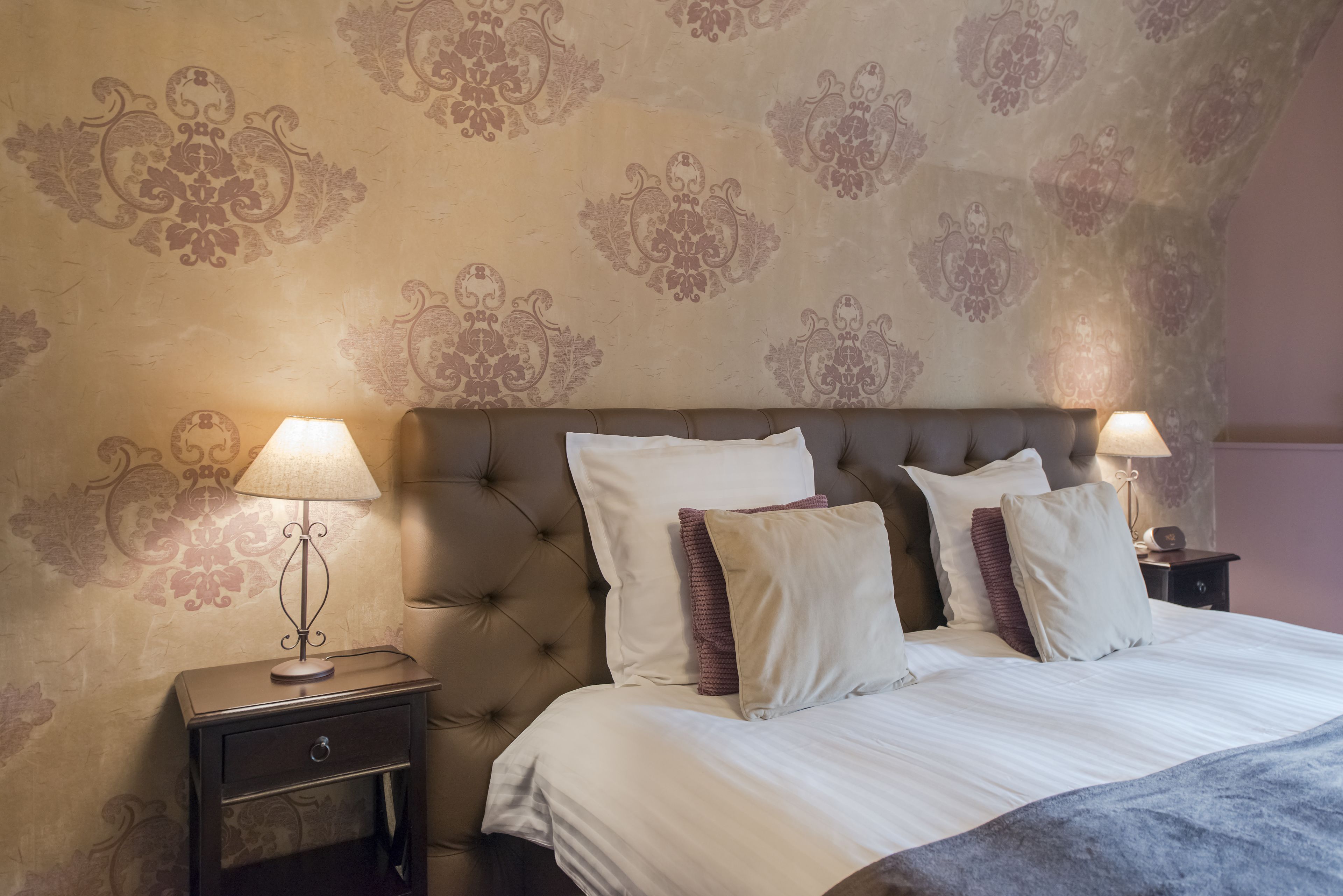 A neatly made bed with white linens and multiple pillows, flanked by two bedside tables with lamps, in a room with patterned wallpaper and a tufted headboard.