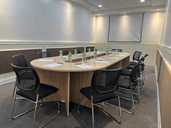 Un salón de conferencias con una larga mesa, sillas negras, botellas de agua y una pantalla para proyector.