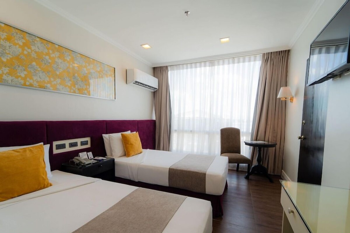 A hotel room with two beds, a desk, a chair, a TV, and a large window with curtains.