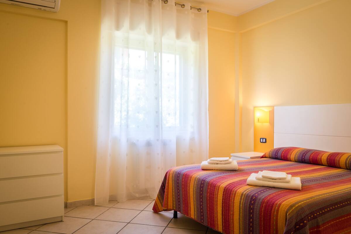 A hotel room with a bed, a bedside table, a lamp, and a window with curtains.