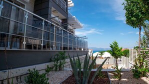 A modern building with a glass balcony, a garden with various plants, and a view of the sea. - Twins Hotel (Cesme)