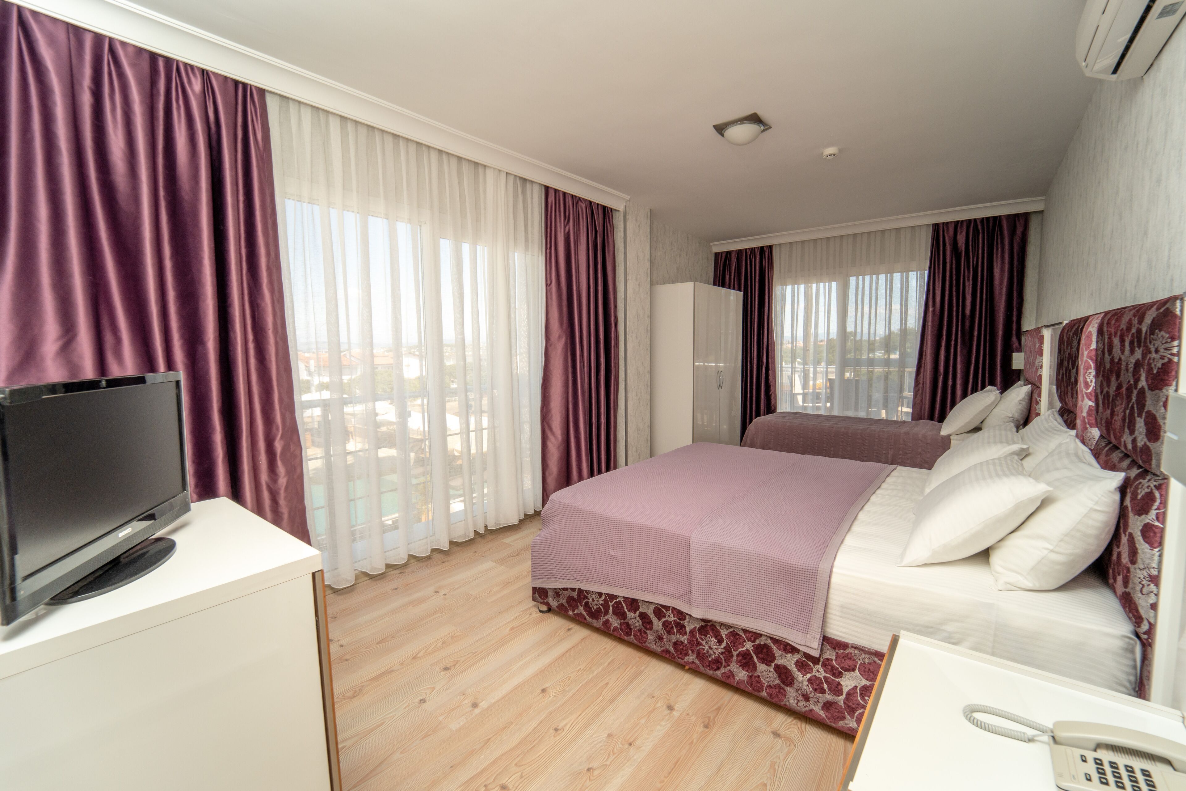 A hotel room with a bed, a TV on a desk, a phone, and a view of the city through large windows.