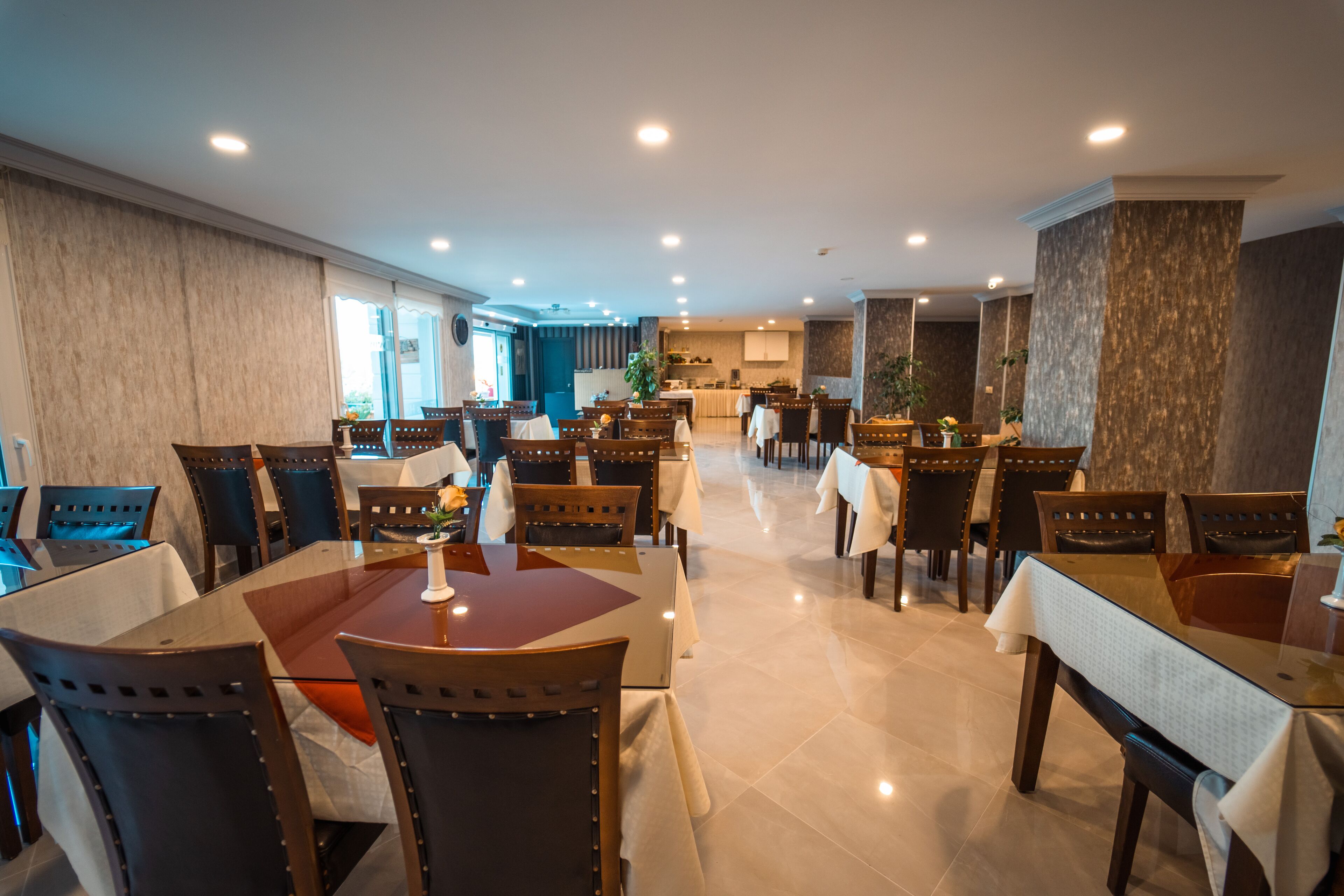 Uma sala de jantar ampla com várias mesas e cadeiras, uma cozinha ao fundo e vista para o exterior.