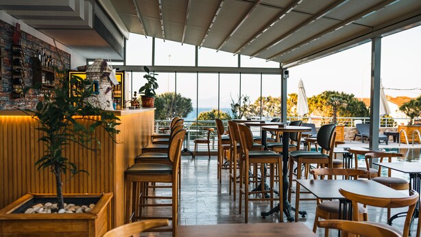 A restaurant with wooden tables and chairs, a bar area with bottles and glasses, and a view of the ocean through large windows. - Twins Hotel (Cesme)