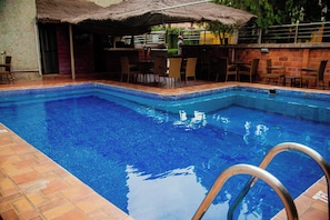 A swimming pool with a tiled edge, a thatched roof structure, and a brick wall. - Denis Hotel (Abuja)