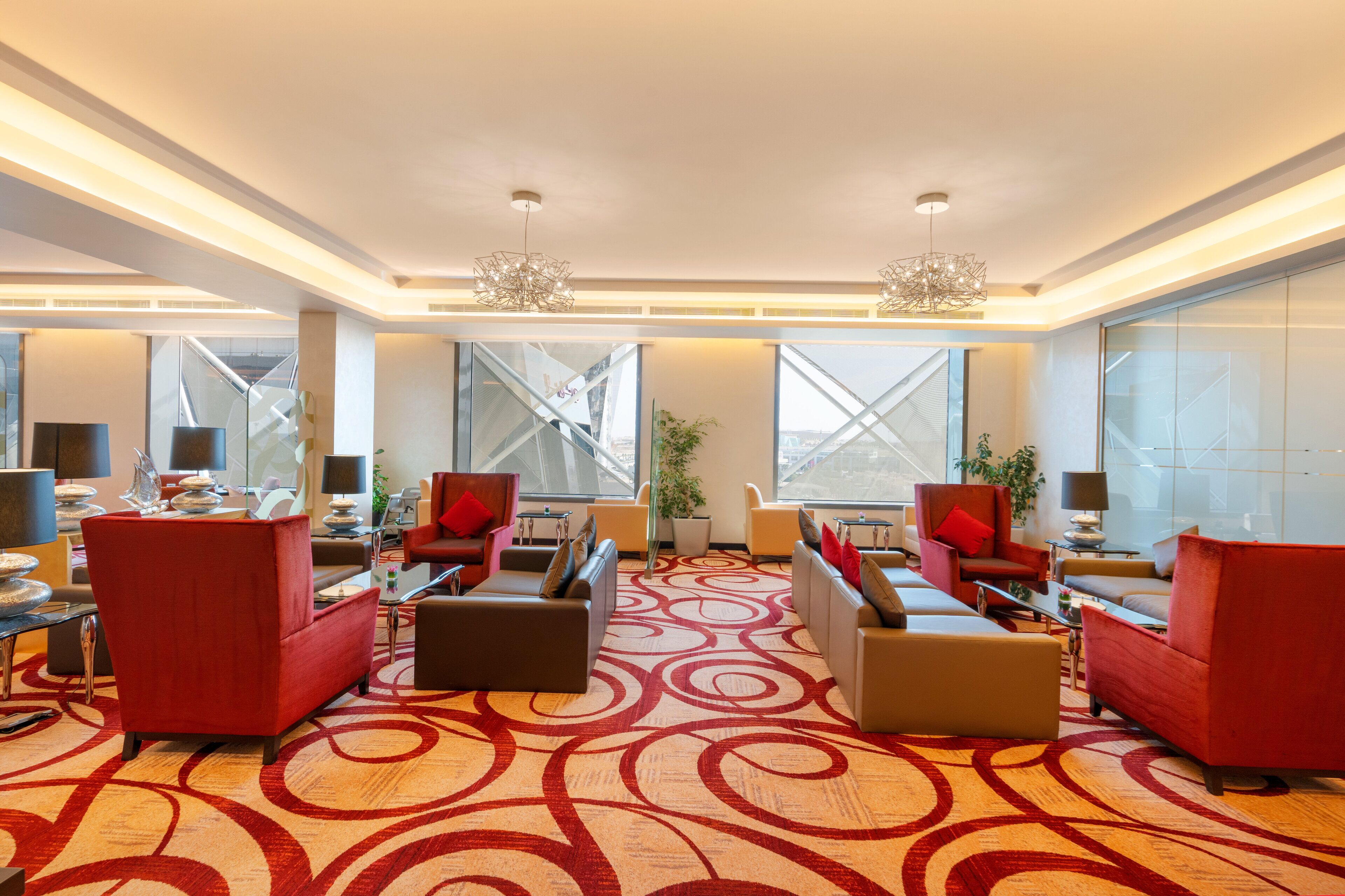 Un hall d’hôtel moderne avec des sièges aux couleurs rouge et beige, de grandes fenêtres et un tapis à motifs.
