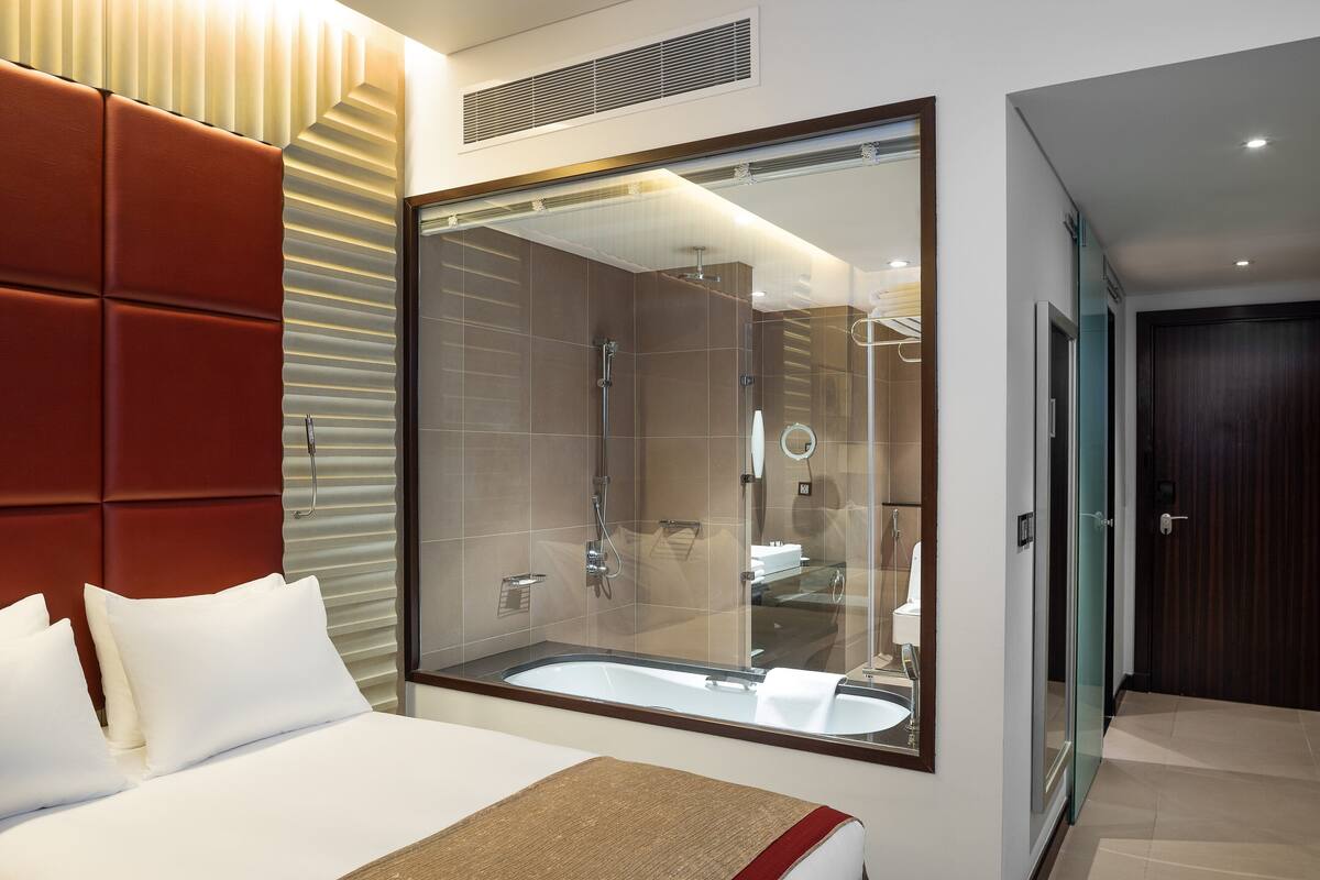 A modern hotel room with a large bed, a glass shower enclosure, and a large mirror reflecting the bathroom.