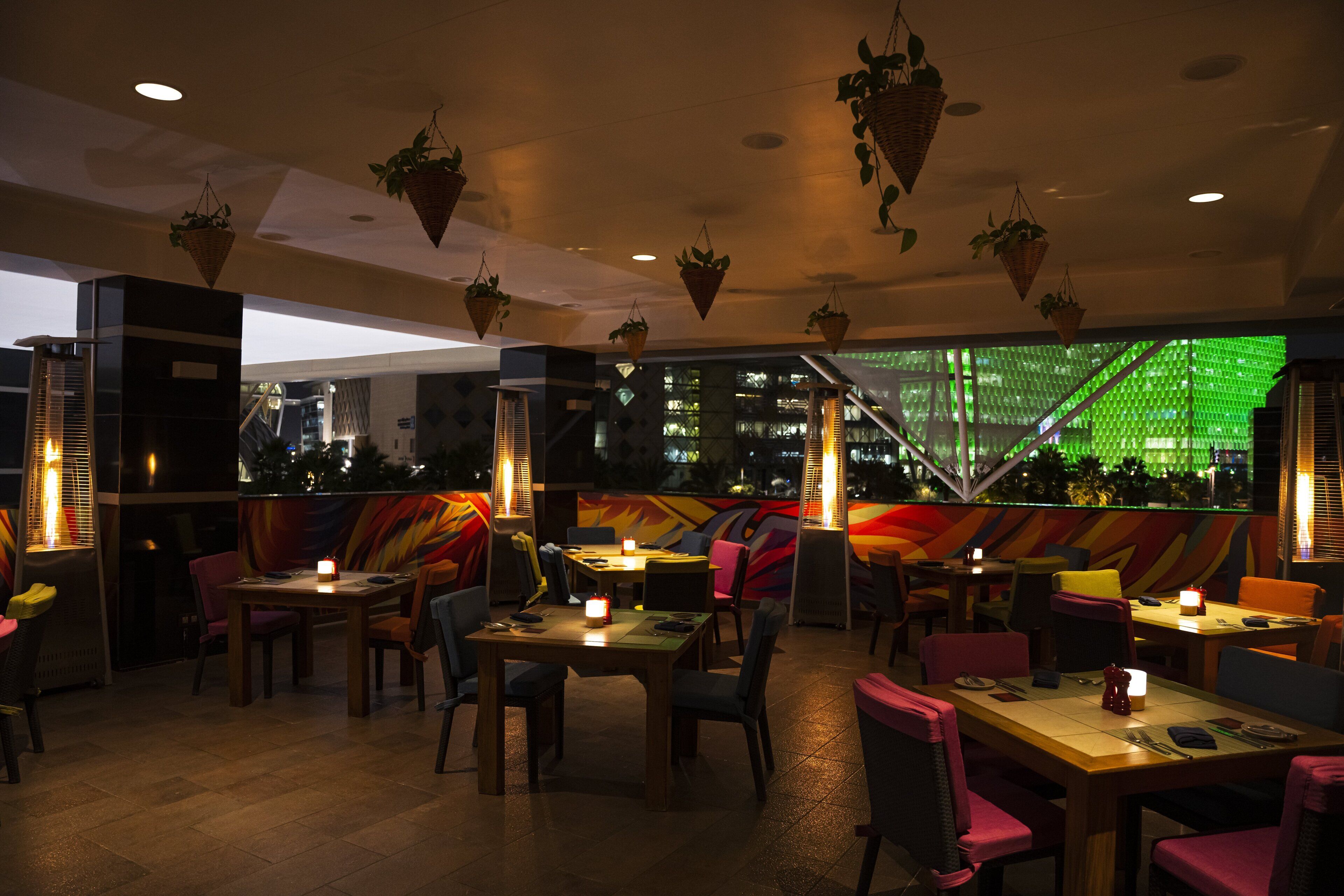 A restaurant with multiple tables and chairs, hanging plants, and a colorful mural on the back wall.