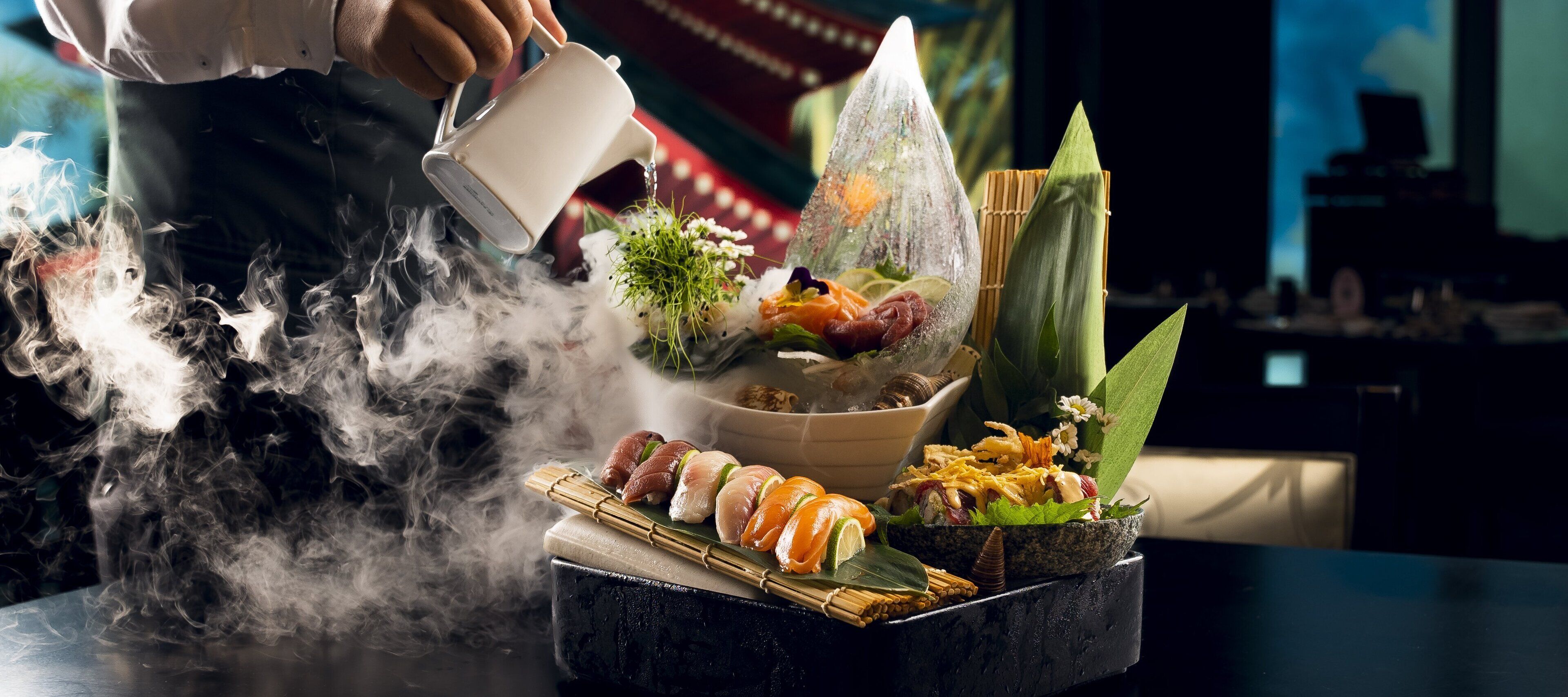A person pouring water onto a table with various food items and smoke.
