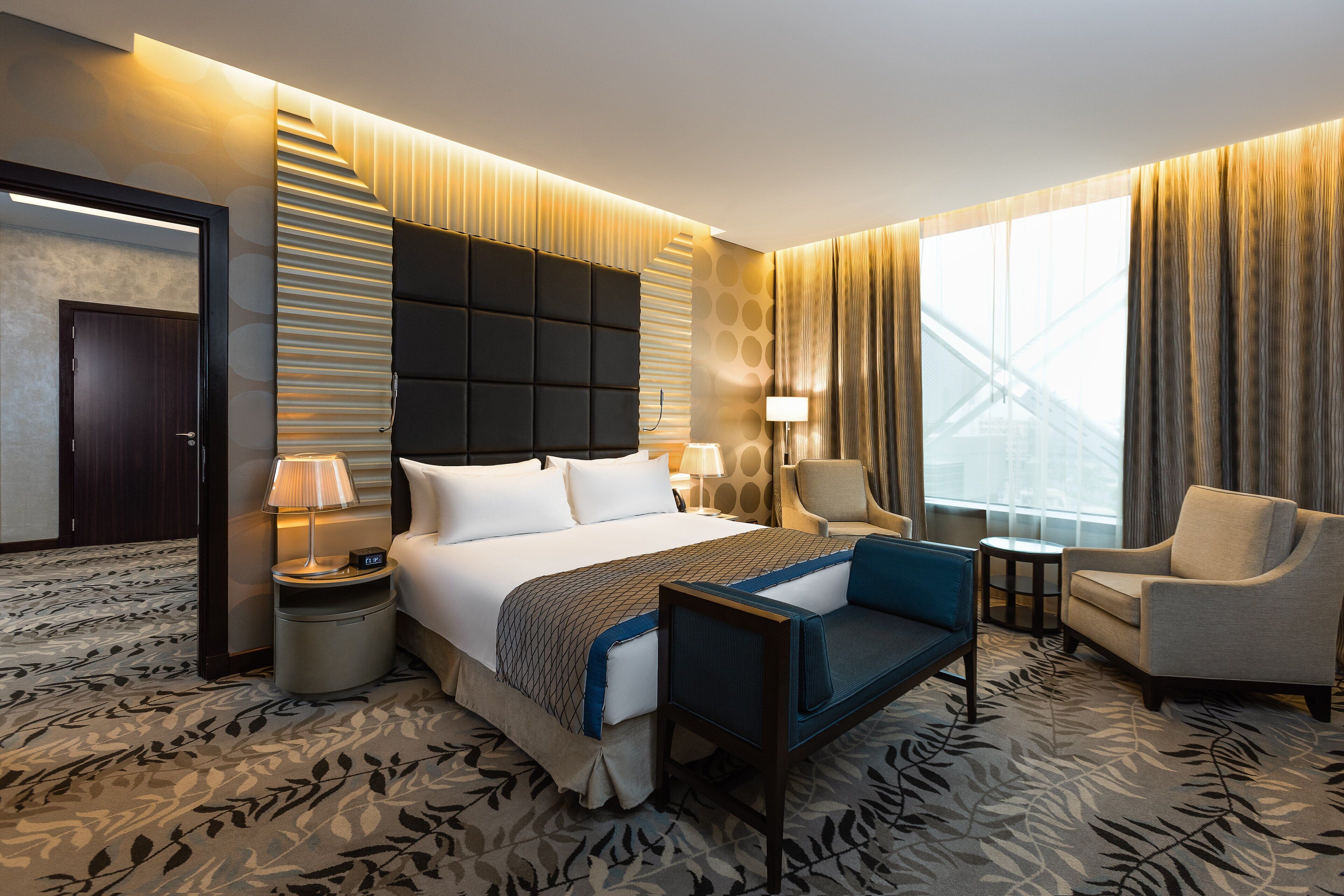 Ein Hotelzimmer mit einem großen Bett, einem Schreibtisch, einem Sessel und Blick auf ein Gebäude durch das Fenster.