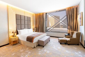 A hotel room with a large bed, a seating area with a chair and ottoman, a nightstand, and a large window with curtains.