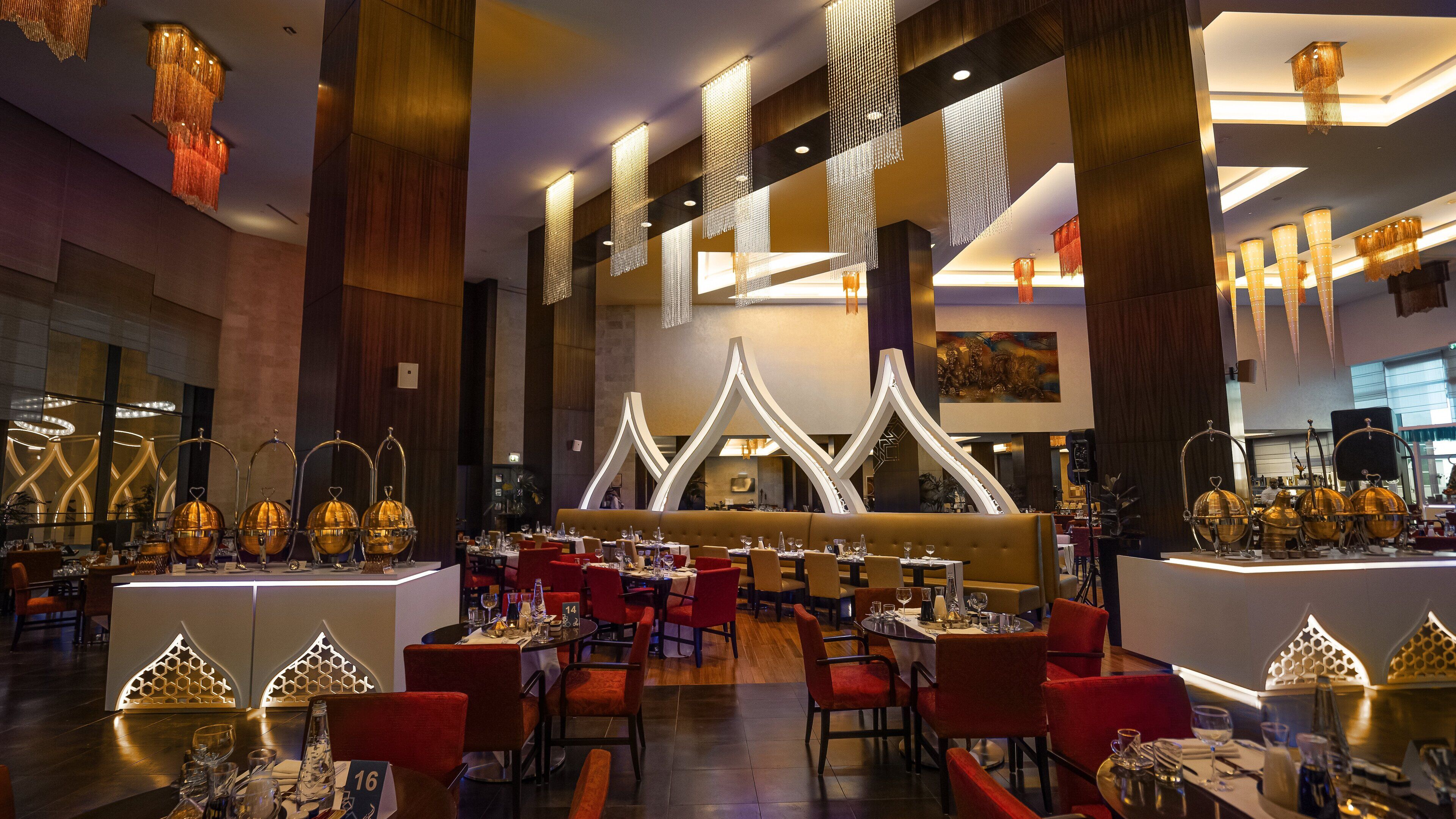 A modern restaurant interior with high ceilings, unique lighting fixtures, and a central seating area with red chairs.