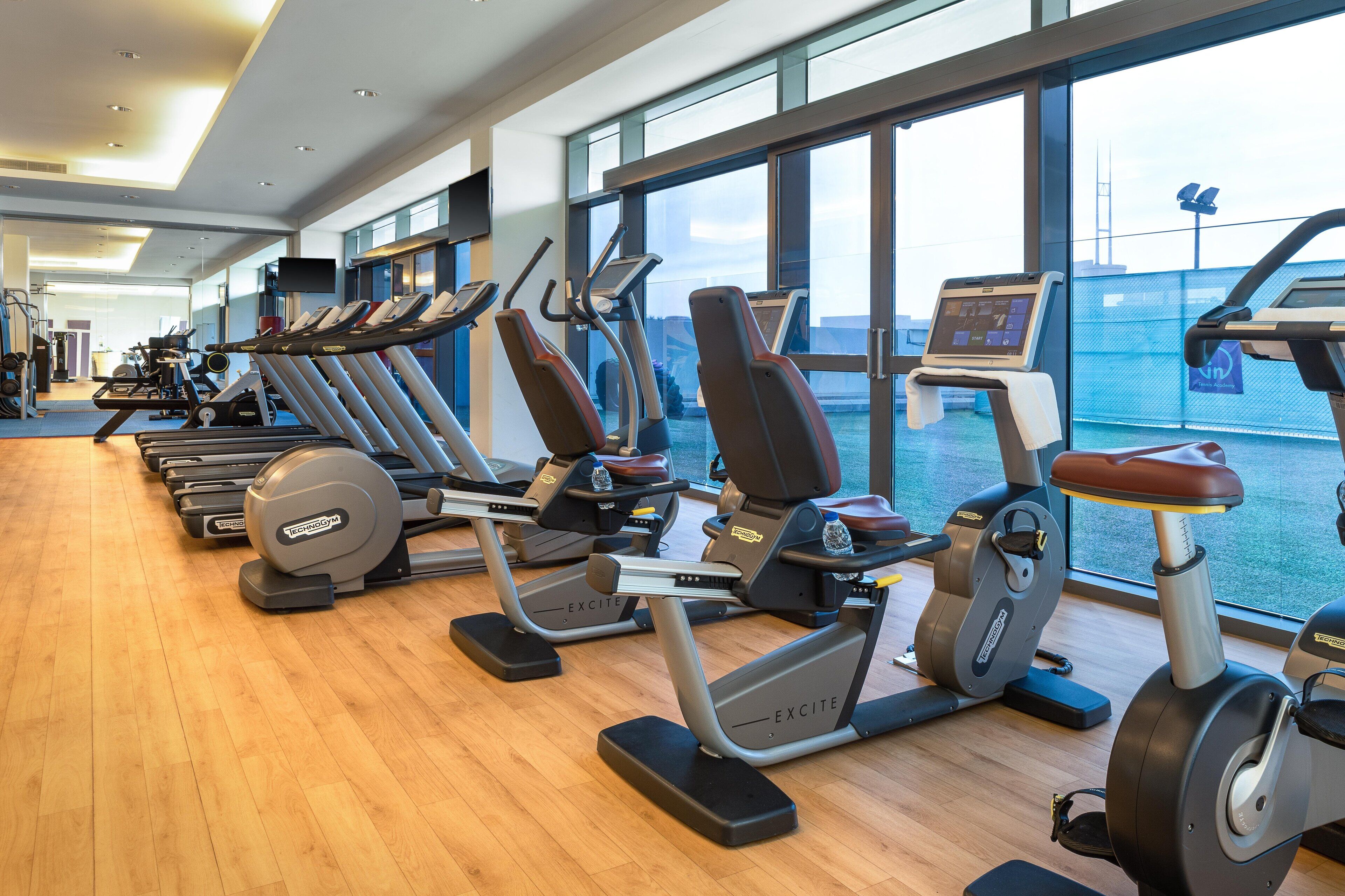 Un gimnasio con varias cintas de correr y bicicletas estáticas, amplias ventanas y suelo de madera.
