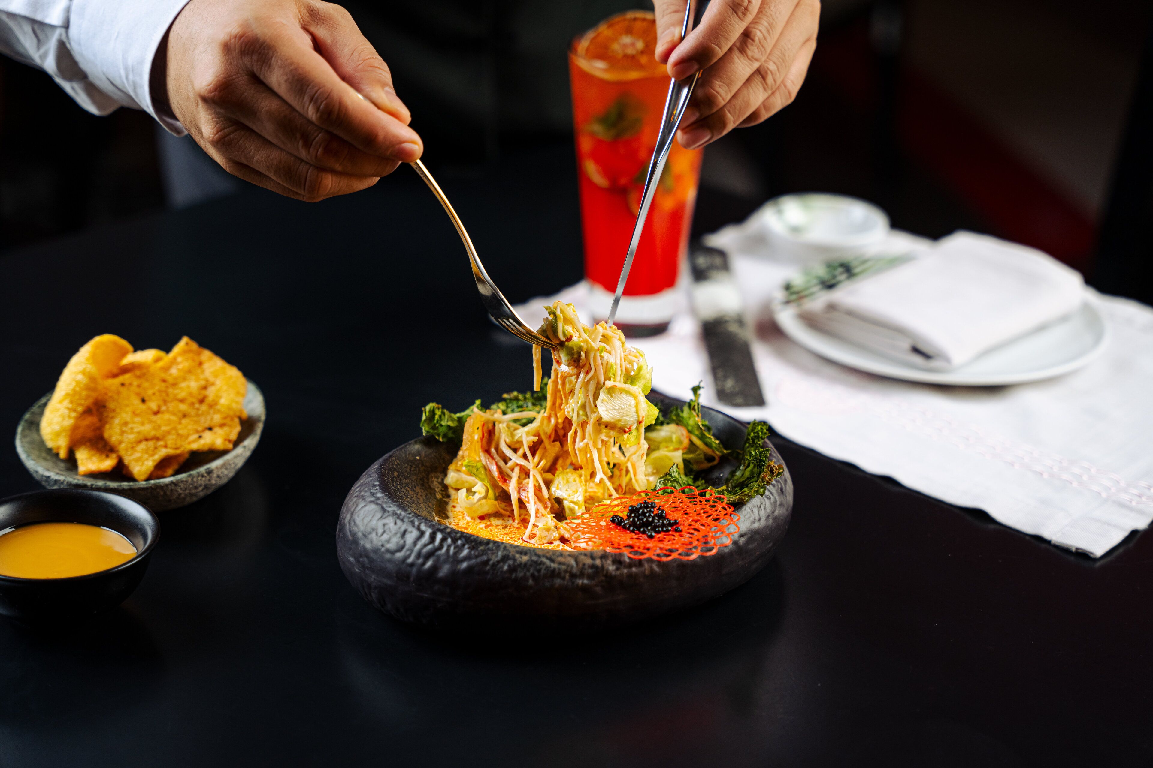 A person eating noodles with chopsticks, a bowl of chips, and a glass of drink on a table.