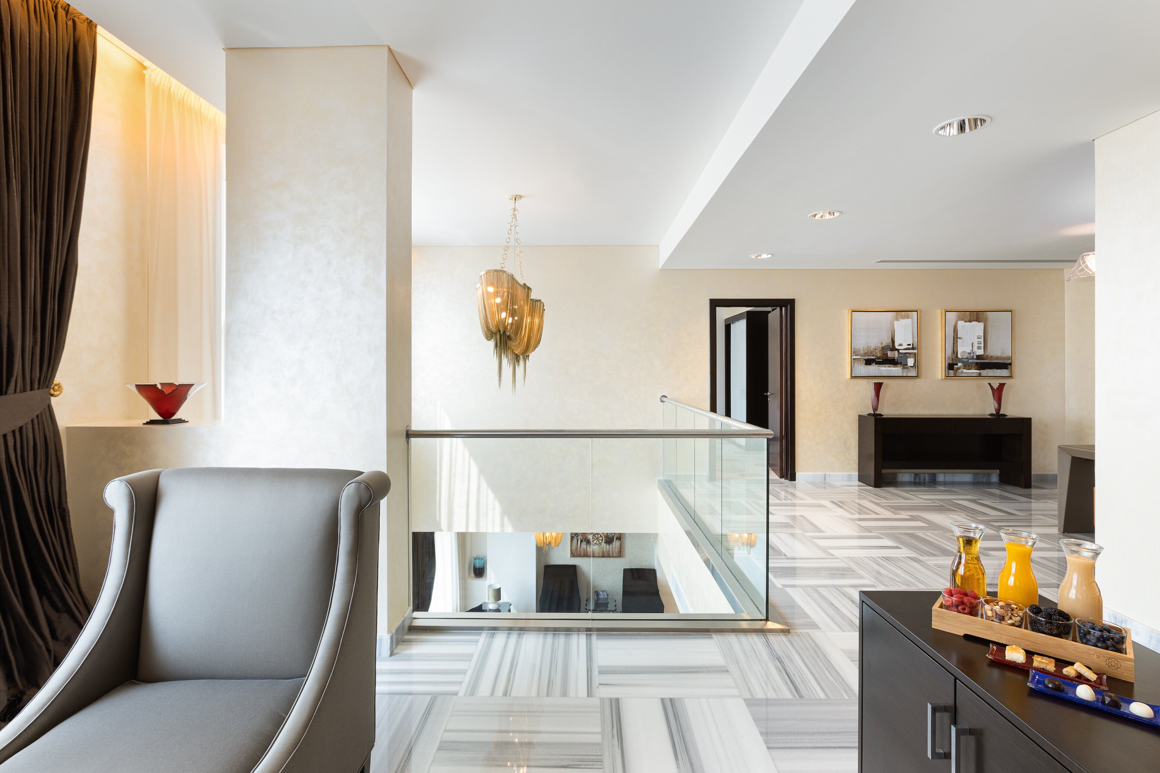 A modern hotel lobby with a marble floor, a grey armchair, a glass railing, and a breakfast tray with fruits and beverages.