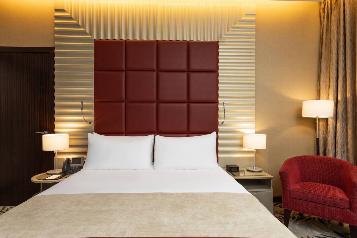 A hotel room with a large bed, two bedside tables, a red armchair, and a headboard with a unique design.
