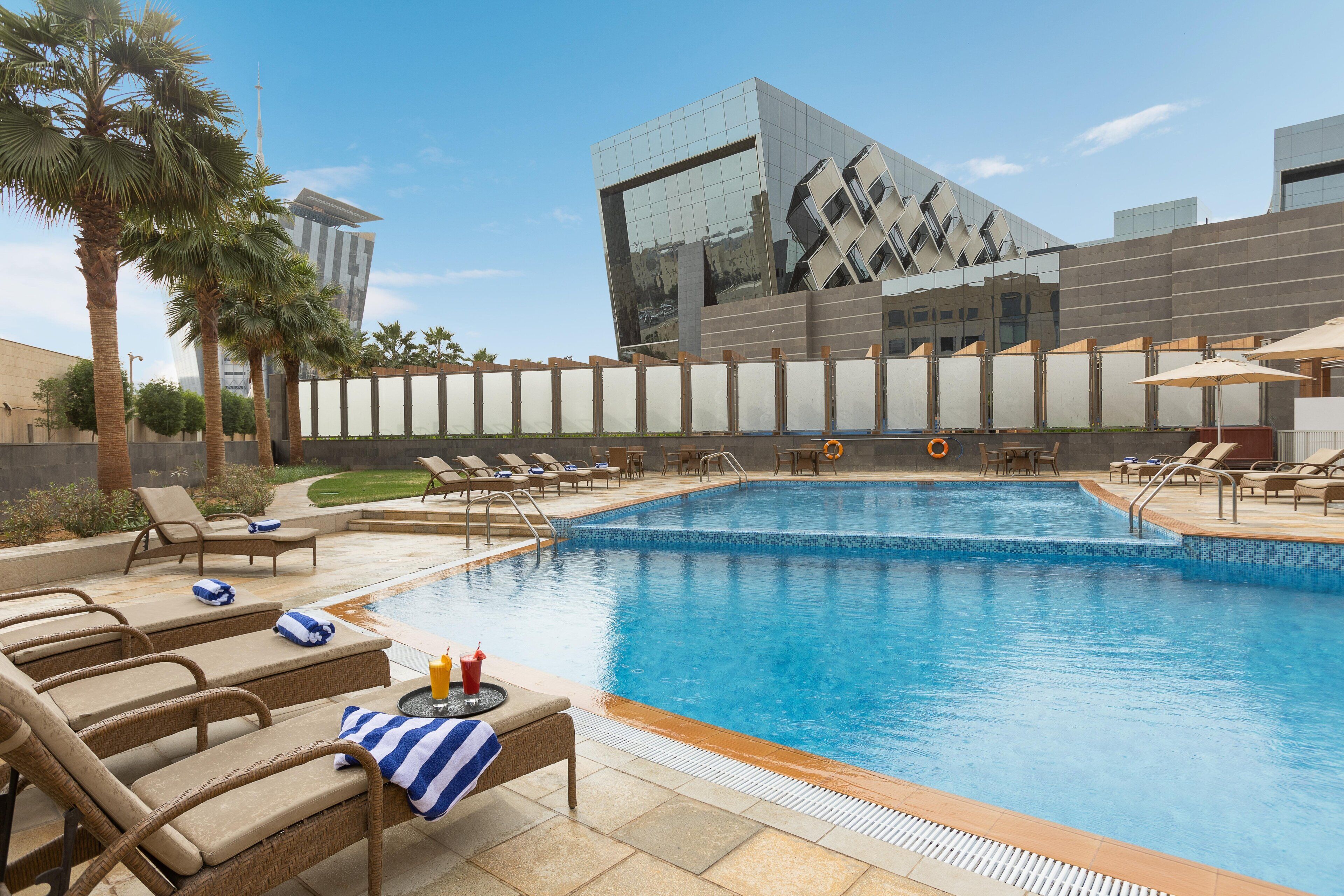 A swimming pool with lounge chairs and umbrellas, surrounded by palm trees and modern buildings.