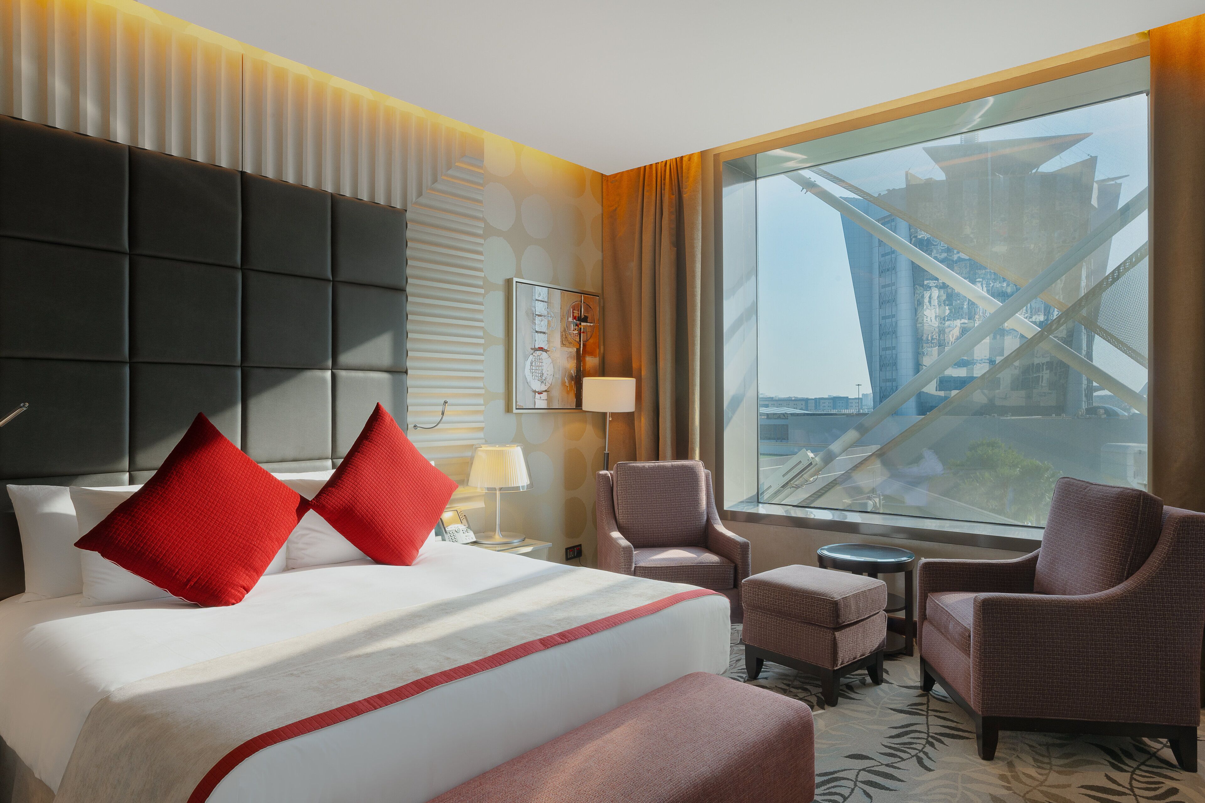 Una camera d'albergo con un letto grande, due cuscini rossi, una sedia, un tavolino e vista su un edificio attraverso una grande finestra.