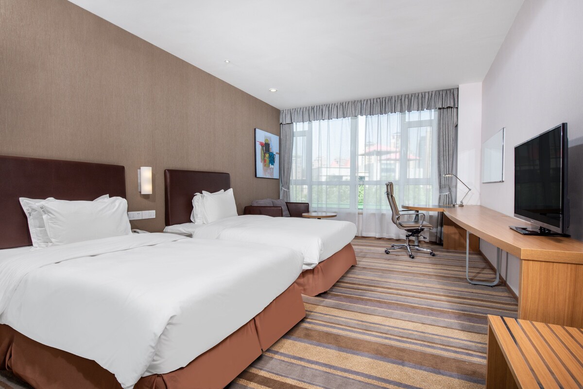 A hotel room with two beds, a desk, a chair, a television, and a large window with curtains.