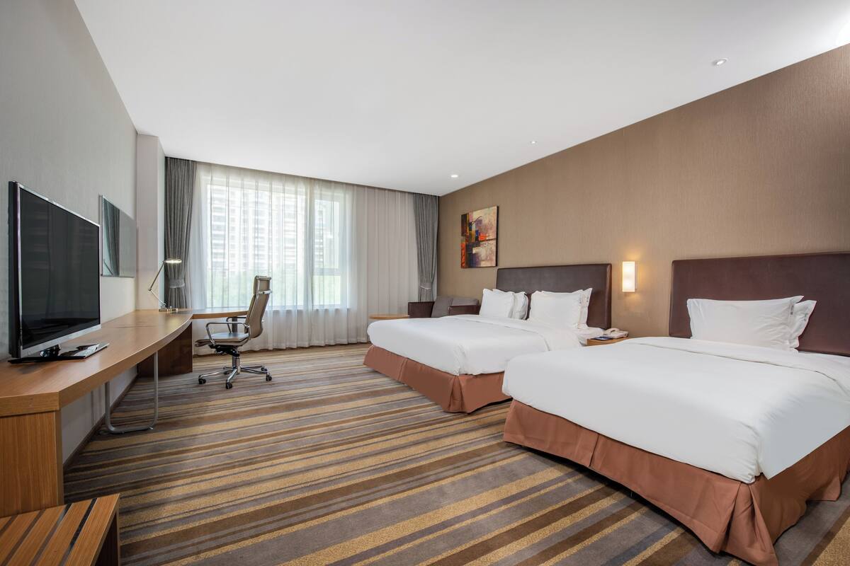 A hotel room with two beds, a desk with a TV, a chair, and a large window with curtains.