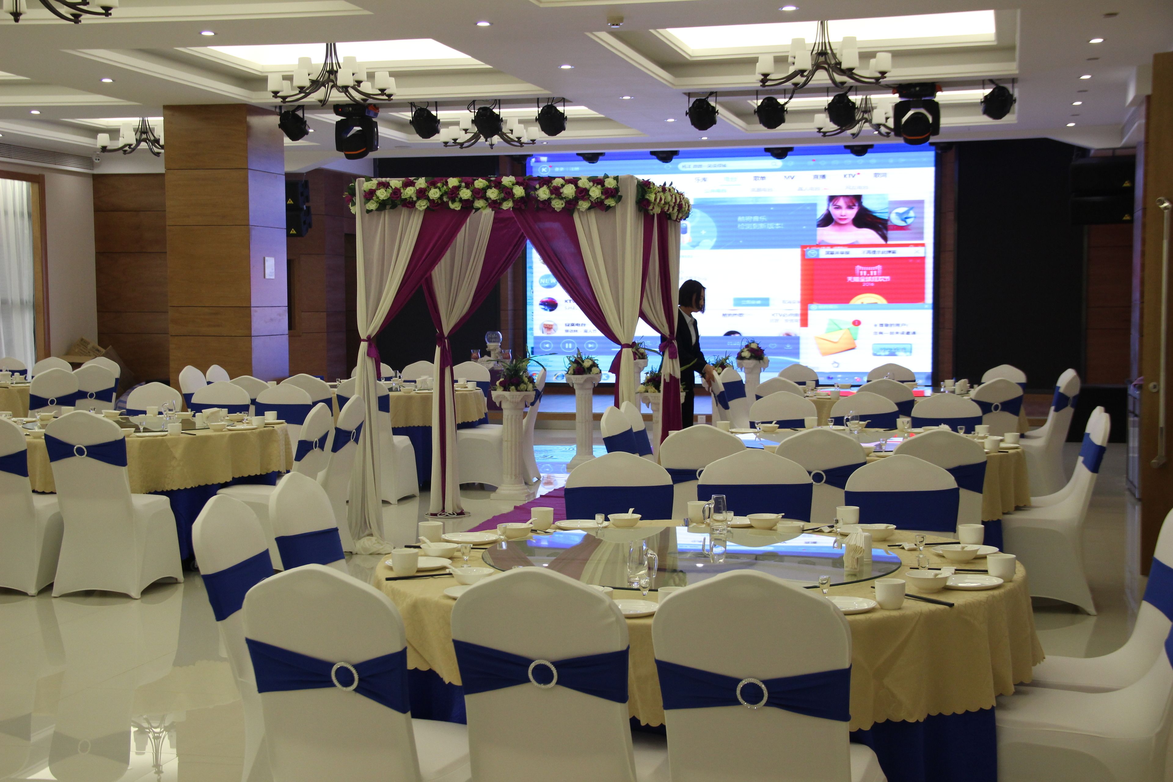 A banquet hall with round tables set for a formal event, featuring white chairs with blue sashes, and a stage with a floral backdrop.