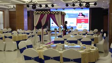 A banquet hall with round tables set for a formal event, featuring white chairs with blue sashes, and a stage with a floral backdrop.