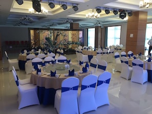 Une salle de banquet avec des tables rondes, des chaises blanches ornées de rubans bleus et un mur décoratif agrémenté de peintures florales.