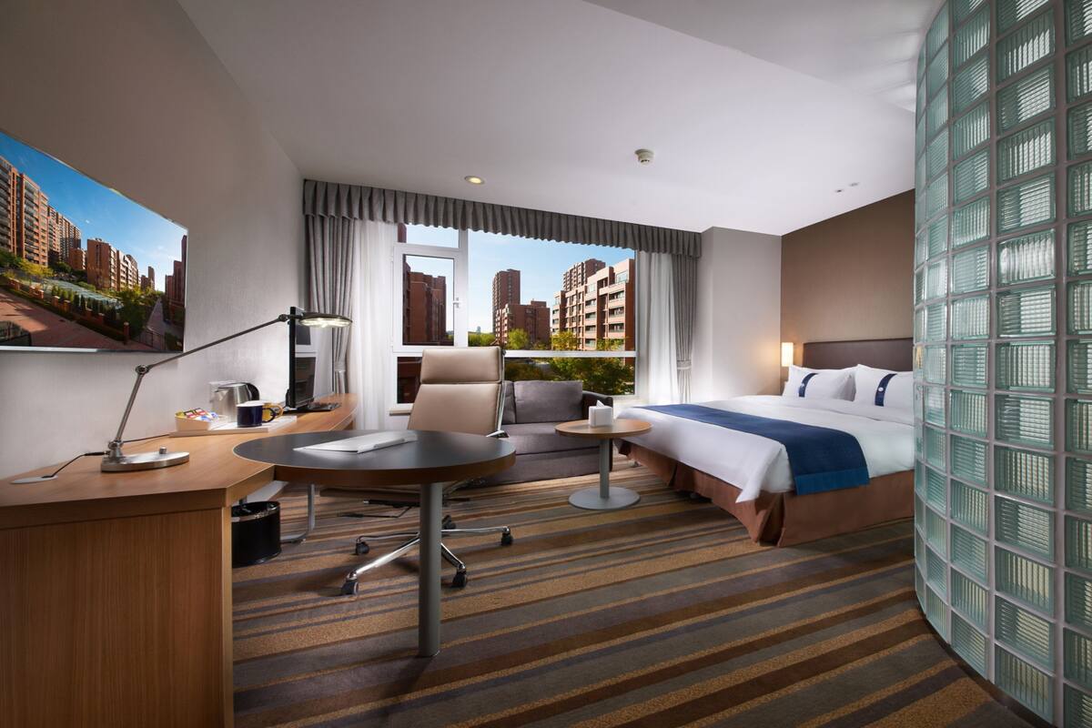 A hotel room with a bed, a desk, a chair, a small table, and a view of the cityscape.