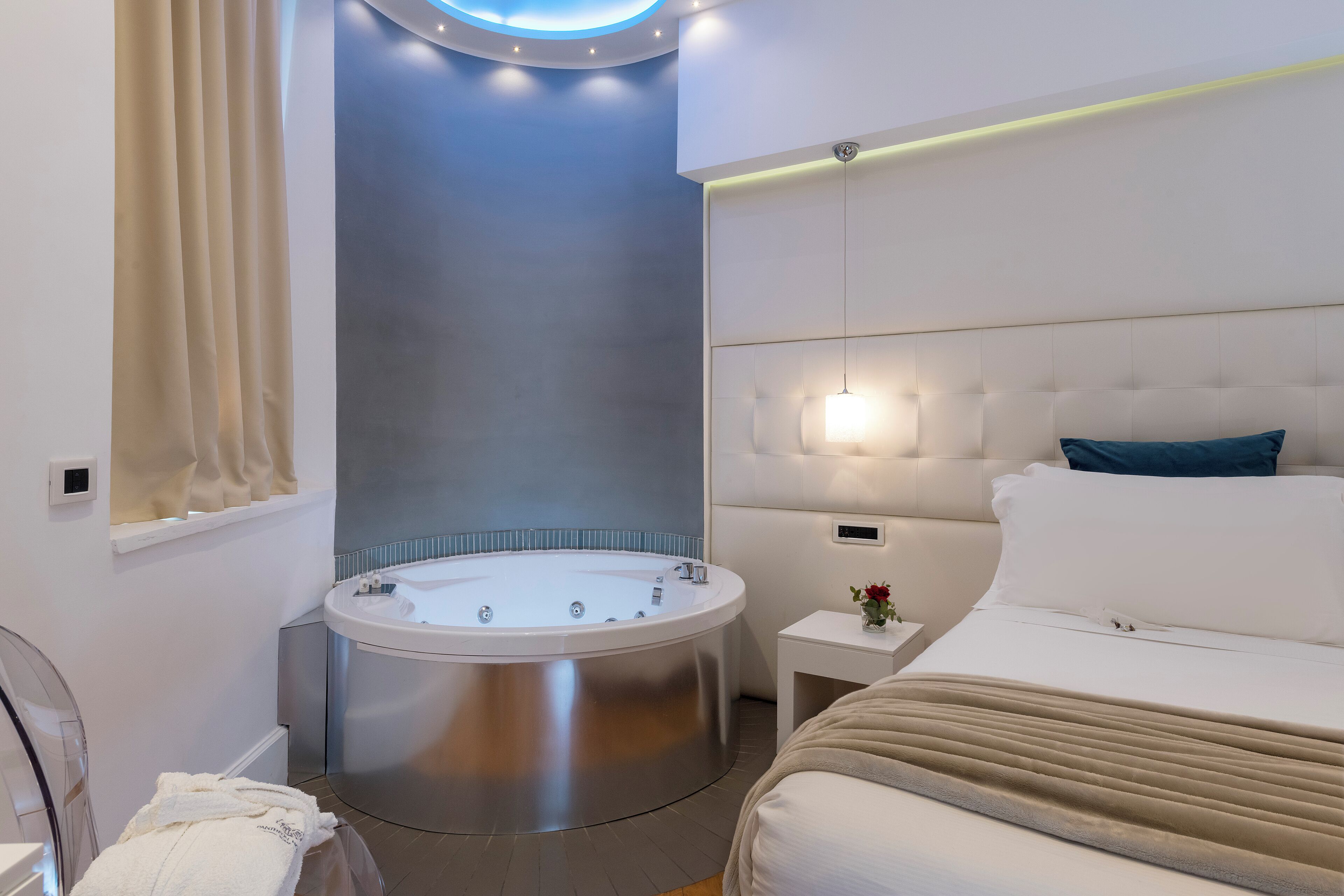 A modern hotel room with a large bed, a round bathtub, and a small bedside table with a lamp.
