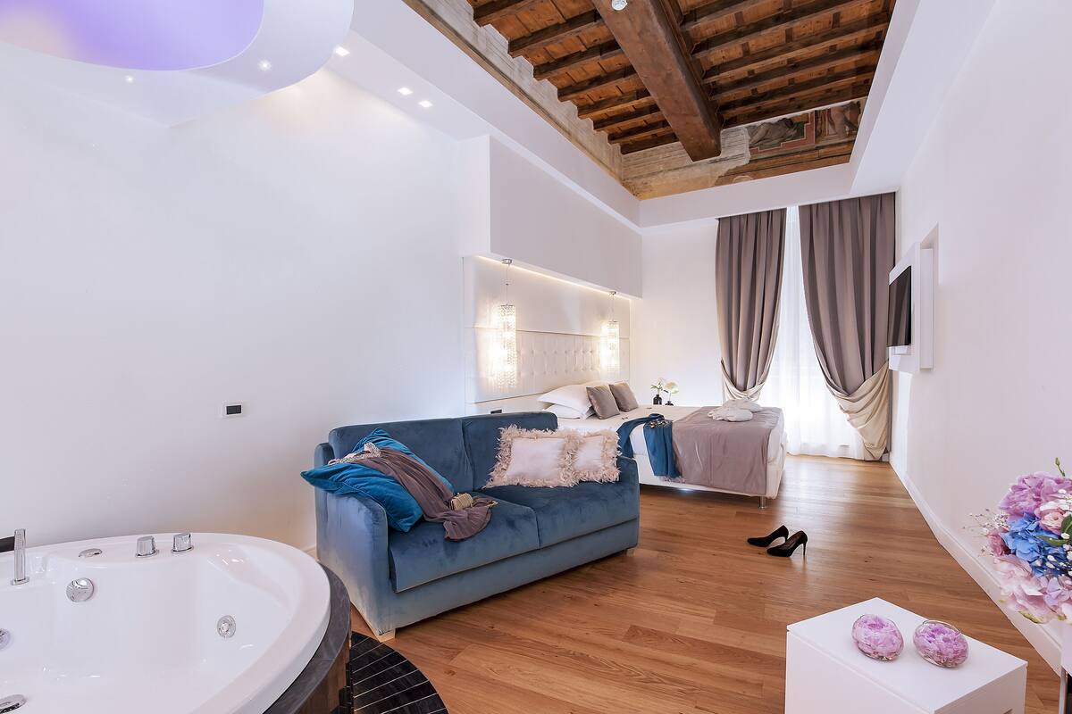 A modern bedroom with a large bed, a blue sofa, a white bathtub, and a wooden ceiling.