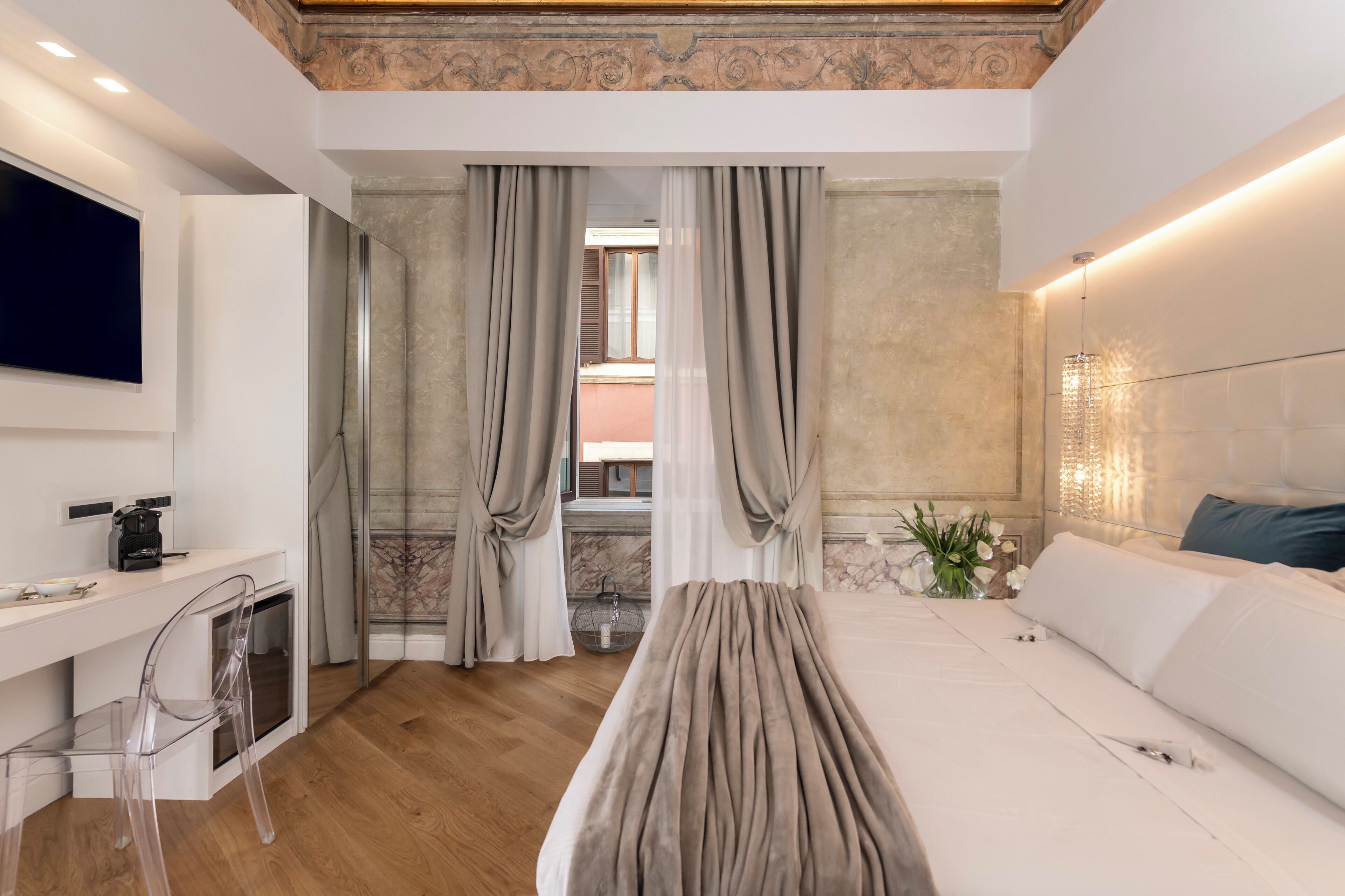 Quarto de hotel moderno com piso de madeira, cama com roupa de cama branca, escrivaninha com cafeteira e televisão montada na parede.