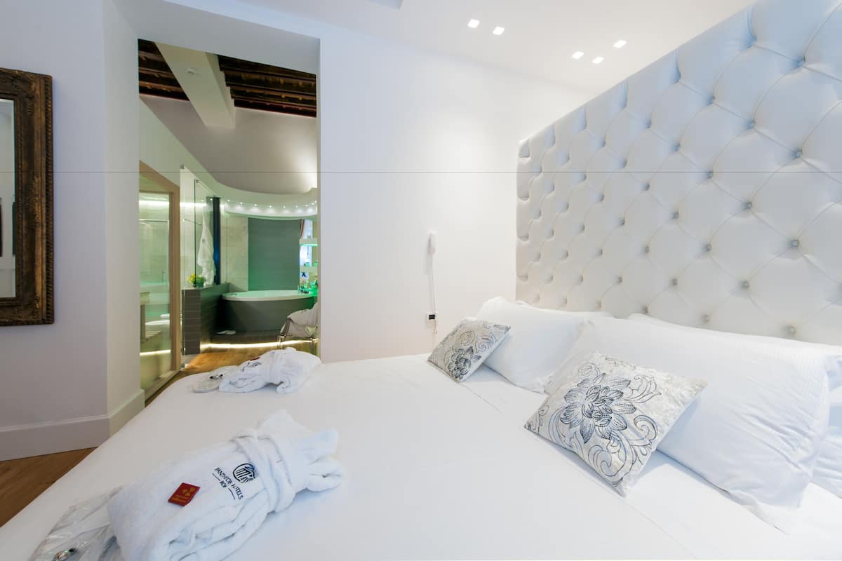 A modern hotel room with a large bed, a tufted headboard, and a bathroom visible through an open door.