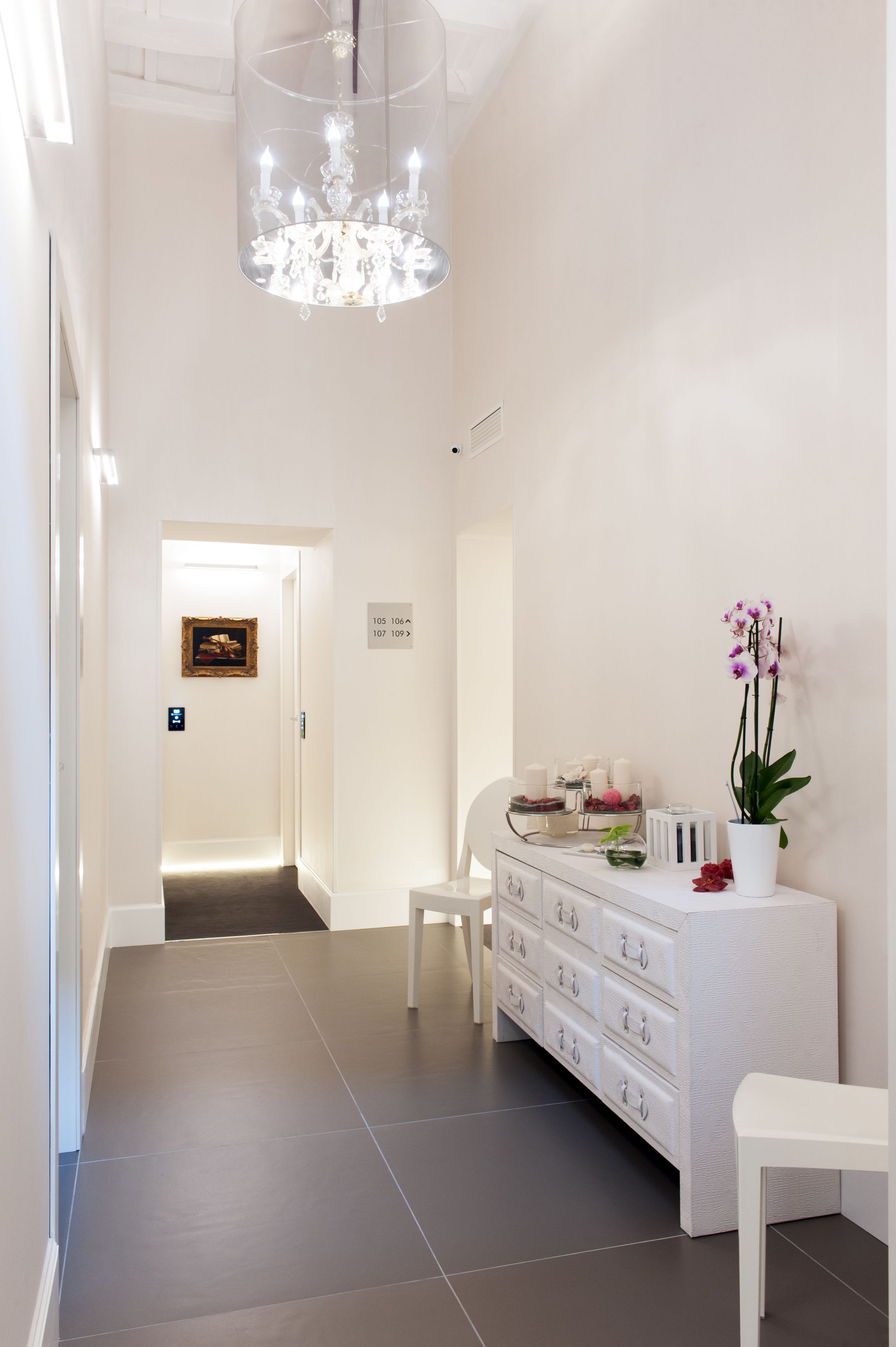 A modern hotel lobby with a chandelier, a reception desk, a white chair, and a potted orchid.