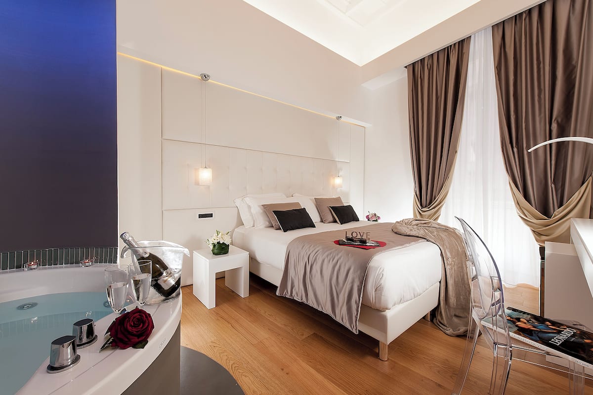 A modern hotel room with a bed, a bedside table, a small table with a bottle and glasses, a chair, and a large window with curtains.
