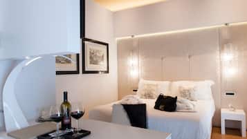 Habitación de hotel moderna con una cama bien hecha, una mesita de noche con una laptop, copas de vino y una botella, y otra mesita de noche más pequeña con una lámpara y un teléfono.