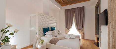 A modern bedroom with a tufted headboard, a flat-screen TV, a wooden floor, and a large window with curtains.