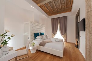 A modern bedroom with a tufted headboard, a flat-screen TV, a wooden floor, and a large window with curtains.