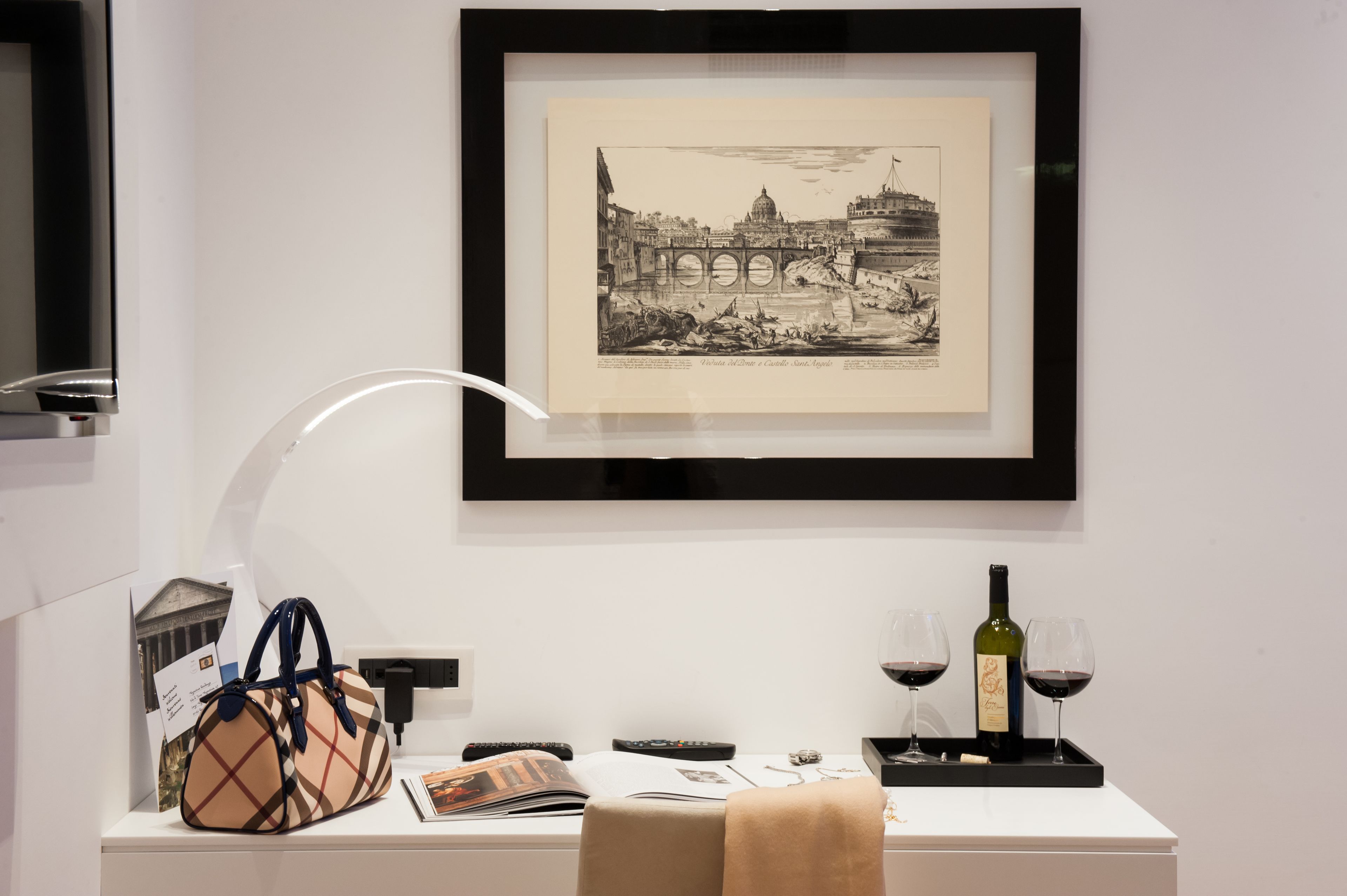 A framed black and white cityscape artwork on a white wall, a beige handbag, a wine bottle, and two wine glasses on a white table.