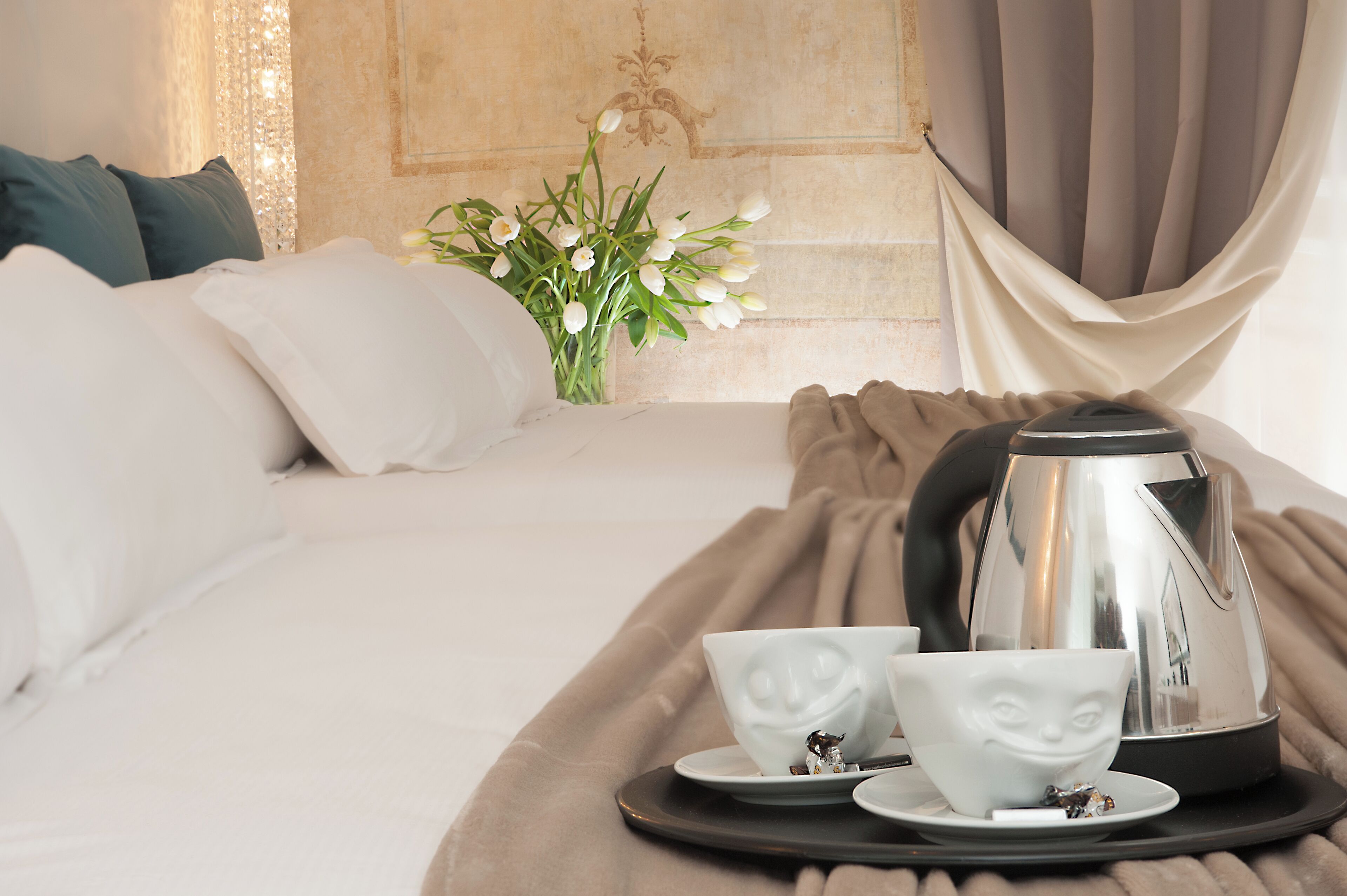 Un letto rifatto con lenzuola bianche, un vaso di fiori bianchi e un vassoio con teiera e tazze.