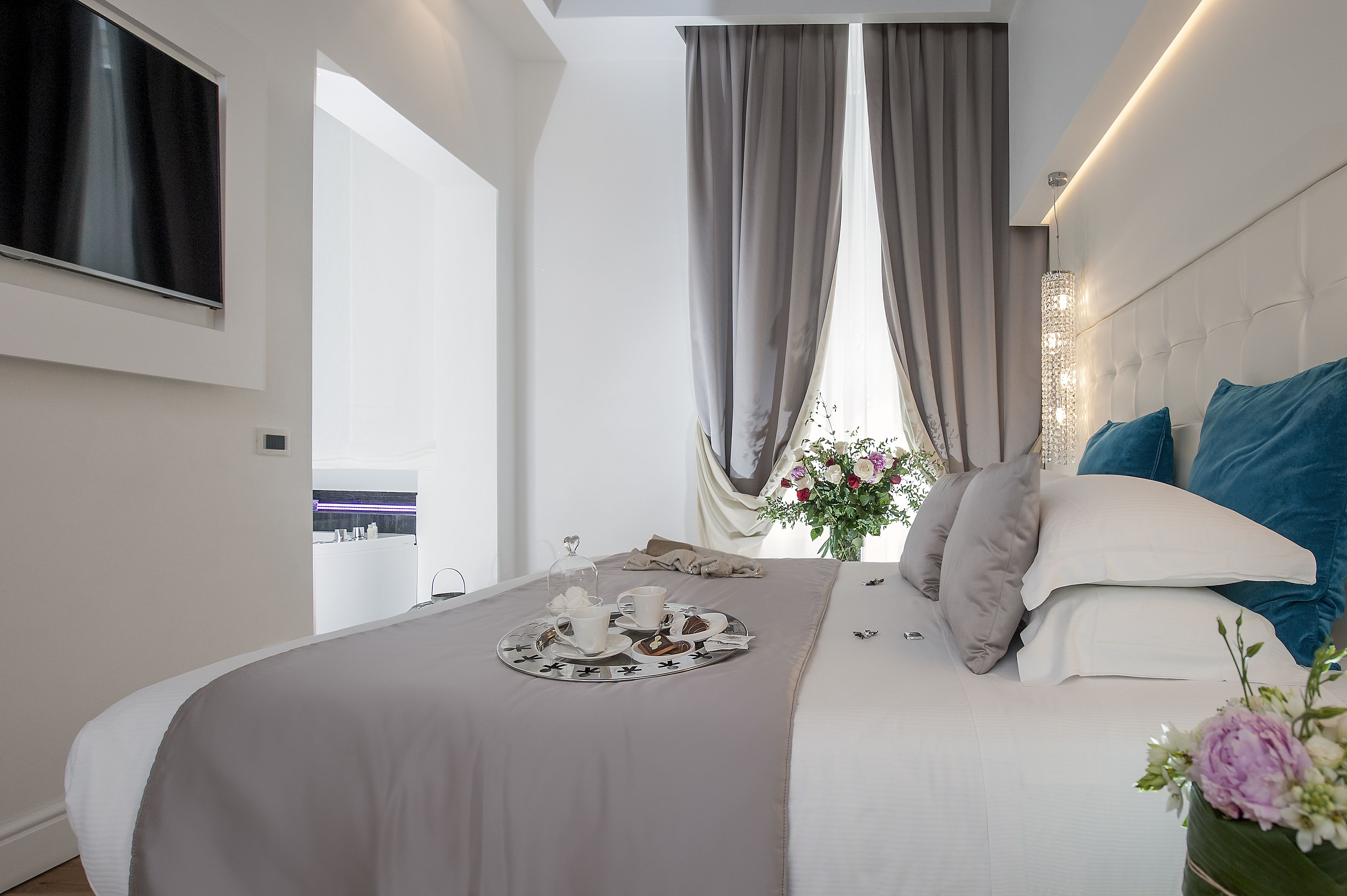 Habitación de hotel moderna con una cama grande, un televisor de pantalla plana, un área de comedor con una bandeja de pasteles y vistas a la ciudad.