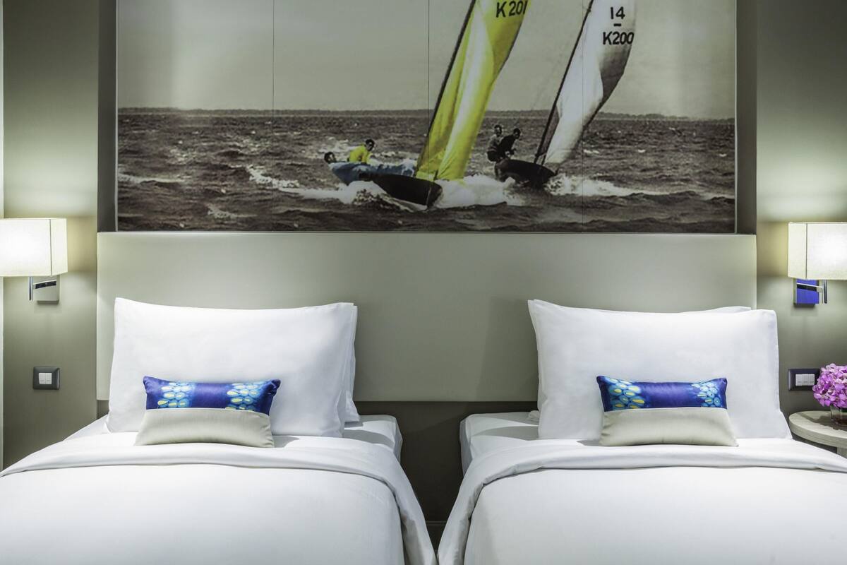 A hotel room with two beds, each with white linens and blue patterned pillows. A large framed photograph of a sailboat on the sea is displayed above the beds.