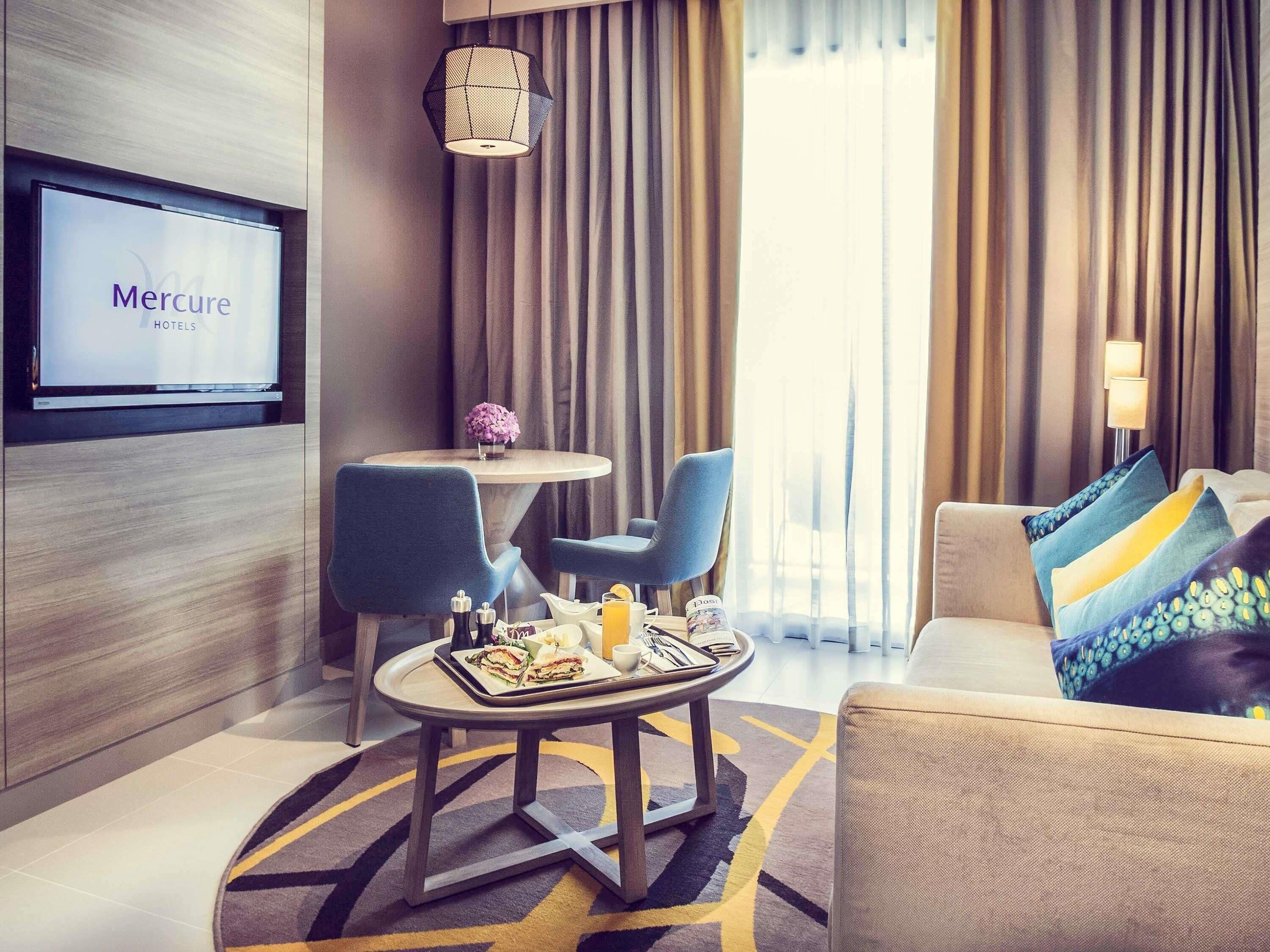 A hotel room with a television displaying 'Mercure HOTELS,' a round table with chairs, a sofa with patterned cushions, and a decorative lamp.