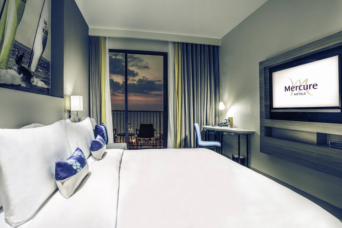 A hotel room with a bed, a desk with a chair, a TV, and a balcony with curtains.