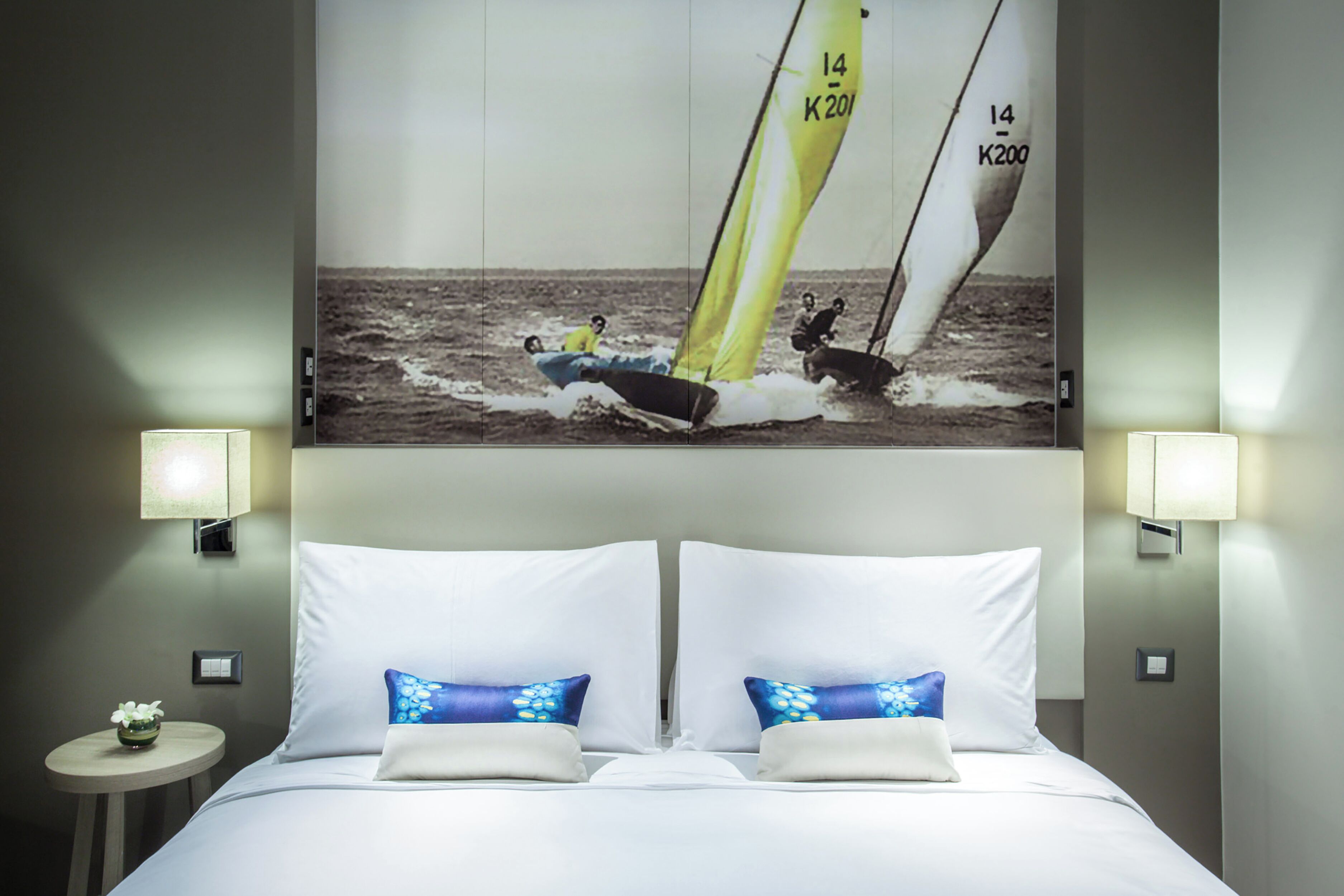 A hotel room with a large sailboat photo on the wall, a bed with white linens, and two bedside lamps.