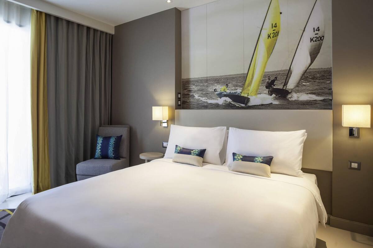 A hotel room with a large bed, two bedside lamps, a chair, and a wall-mounted painting of sailboats.